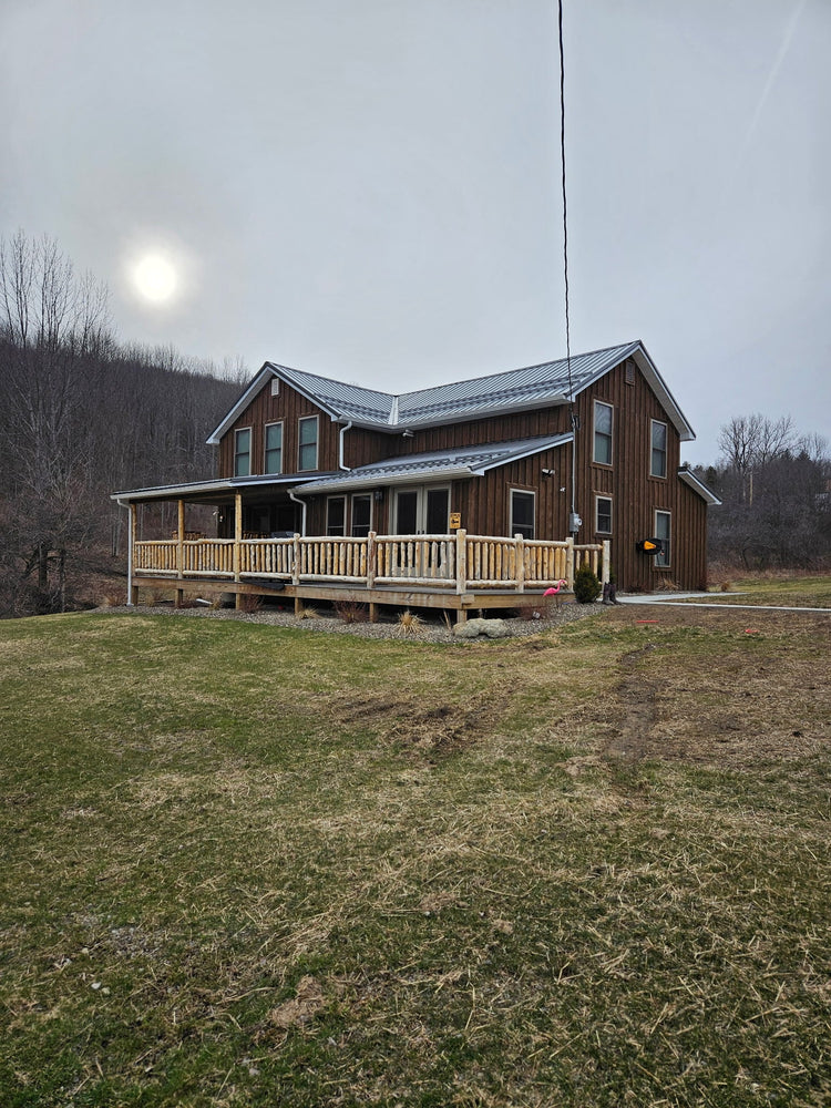 "Rustic two-story wooden cabin with metal roof and wraparound porch on grassy hillside in Steuben County, NY, under a cloudy sky."