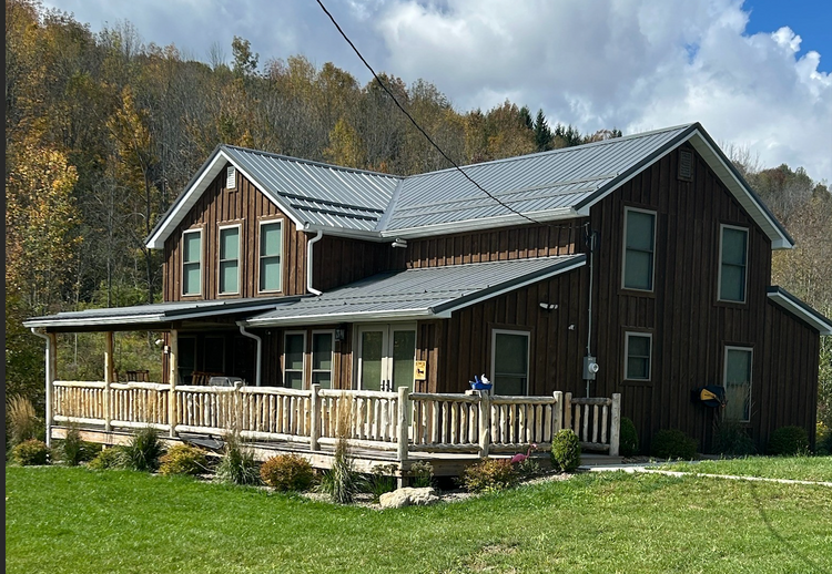 "Rustic two-story wooden cabin with metal roof and wraparound porch on grassy hillside in Steuben County, NY, under a cloudy sky."